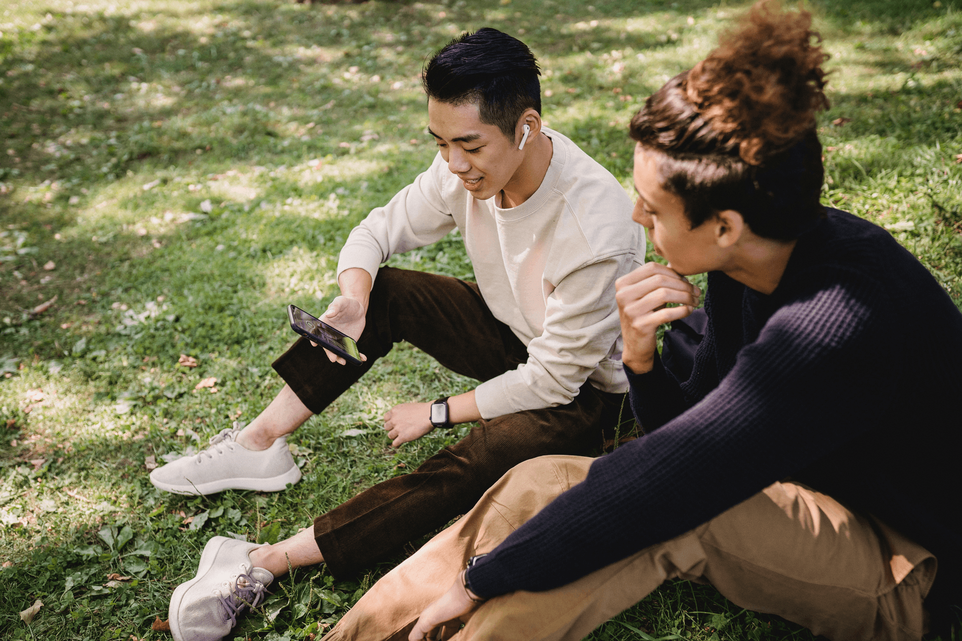 two men looking at a mobile phone while sitting on a grass field