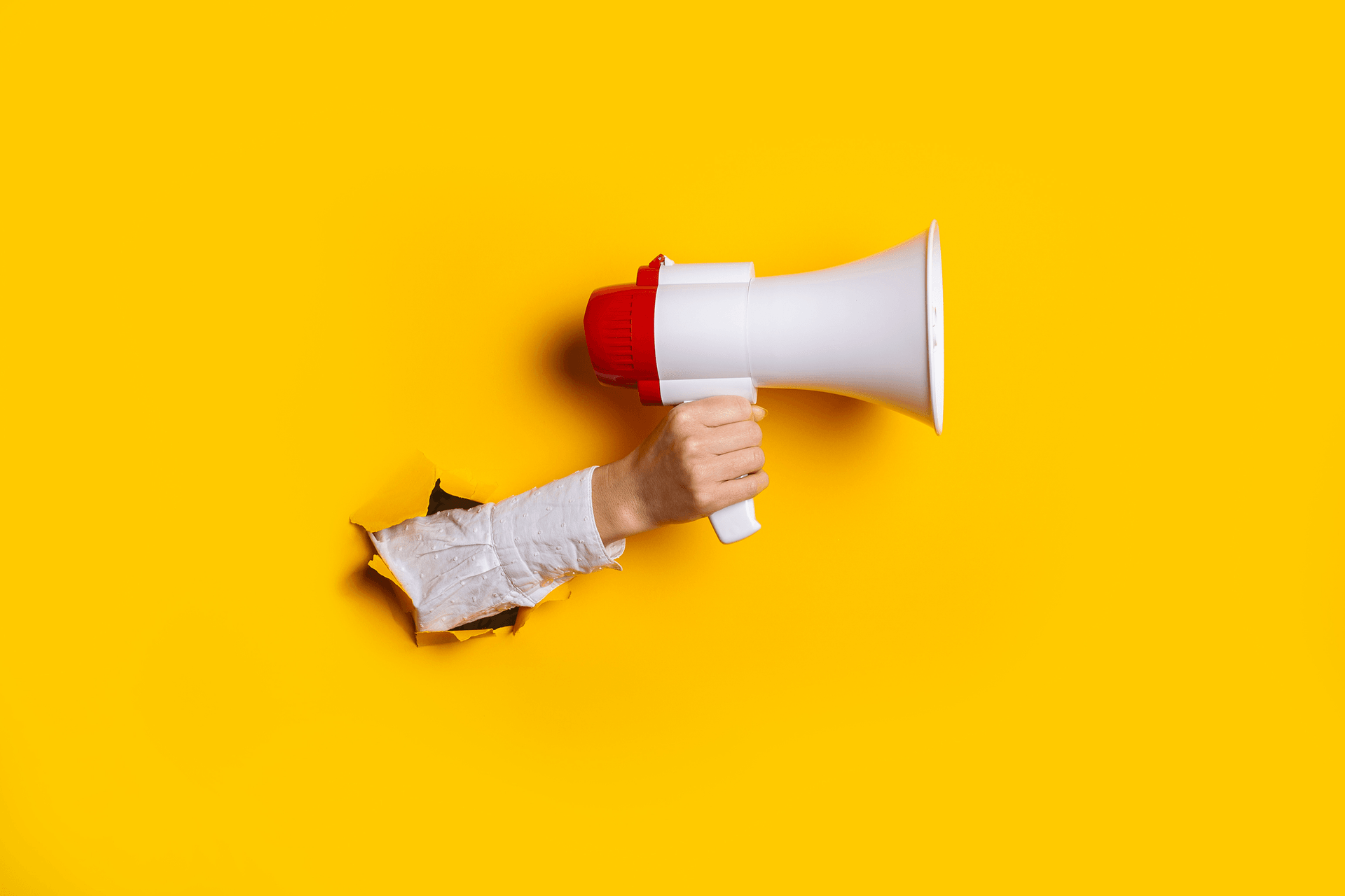 Hand holds a megaphone from a hole in the wall on a yellow background