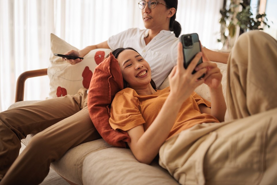 Woman watching a video on her phone while sitting with a man watching tv.